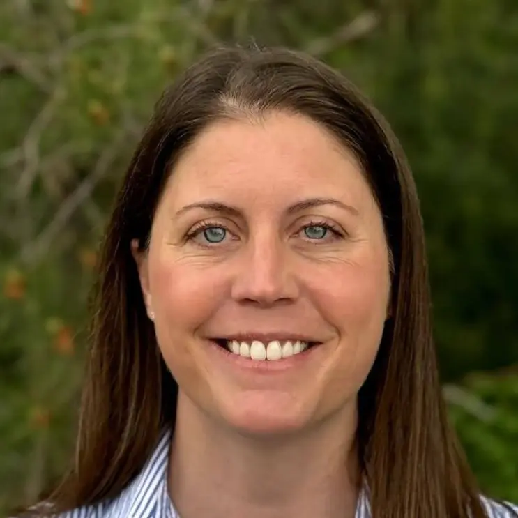 A woman smiling outdoors with greenery in the background.
