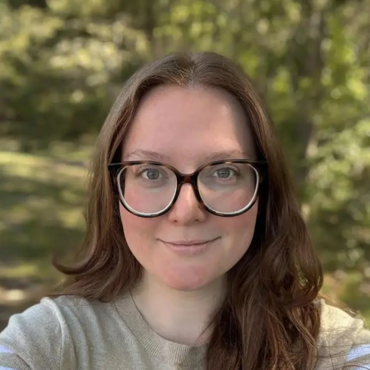 A young woman with glasses smiling outdoors in a natural setting.